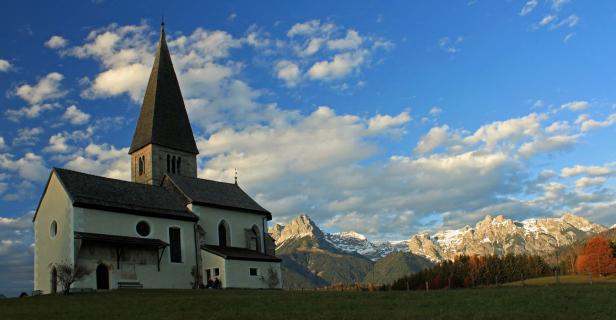 Eine Kirche mit hohem Turm vor einer Bergkulisse unter blauem Himmel mit Wolken.