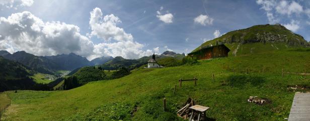 Panorama einer grünen Berglandschaft mit einer kleinen Kirche und einem Bauernhaus.