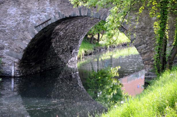 Eine Steinbrücke überspannt einen kleinen Fluss, dessen Oberfläche das Grün der Ufervegetation reflektiert.