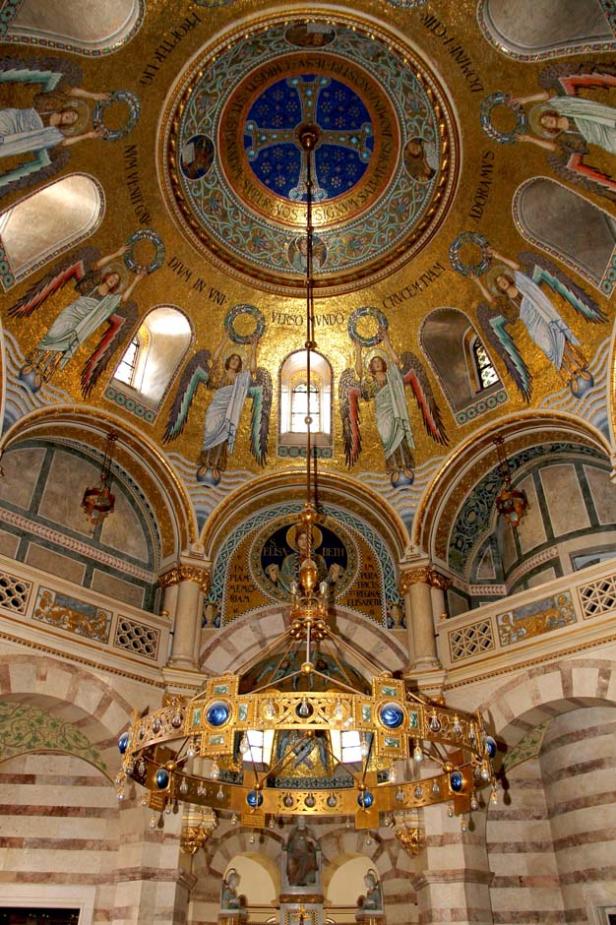 Innenansicht einer Kirche mit einem goldenen Kronleuchter und Mosaiken an der Decke.