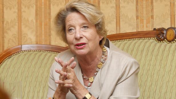 Eine ältere Frau mit blonden Haaren sitzt auf einem Sofa und spricht.