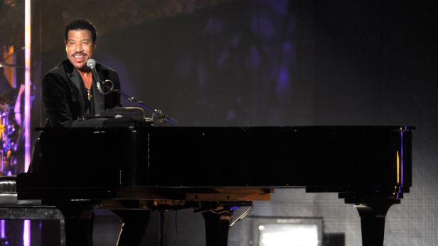 Lionel Richie spielt auf einem schwarzen Klavier und singt in ein Mikrofon.