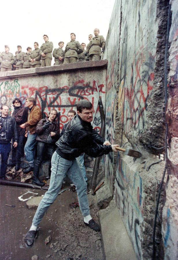 Ein Mann schlägt mit einem Hammer auf die Berliner Mauer ein, während Soldaten zusehen.
