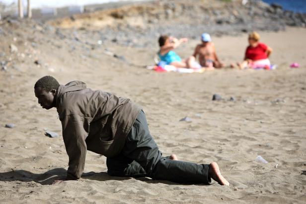 Ein Mann krabbelt barfuß am Strand, während sich im Hintergrund Badegäste sonnen.