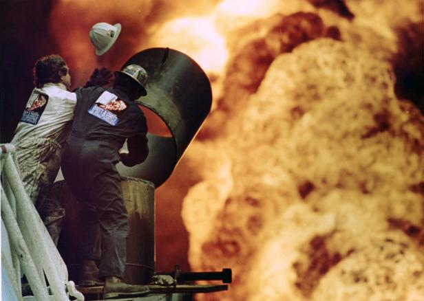 Zwei Arbeiter stehen vor einer riesigen Feuerwand, einer wirft seinen Helm in die Luft.