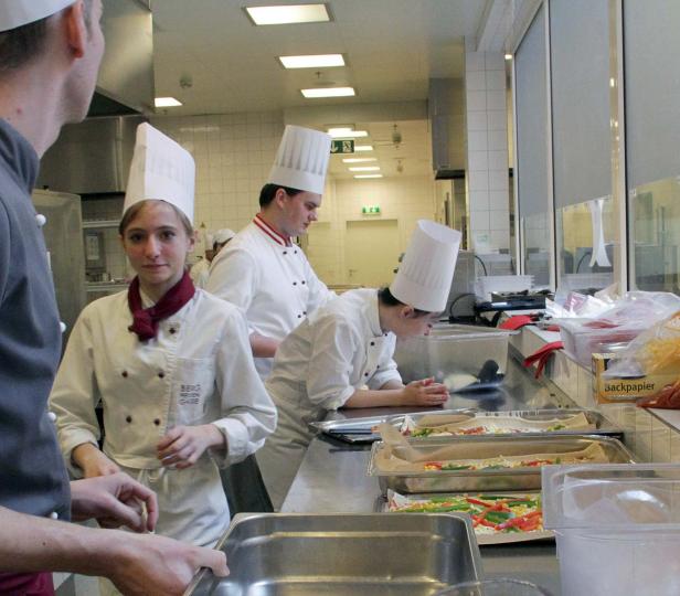 Mehrere Köche bereiten in einer Großküche Speisen zu.