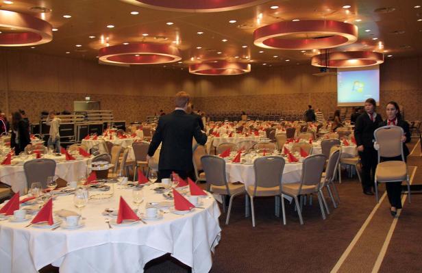 Ein großer Saal ist für eine Veranstaltung mit runden Tischen und roter Dekoration vorbereitet.