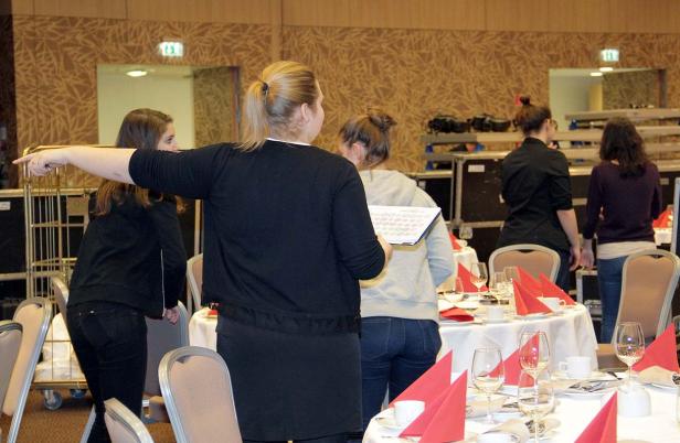 Eine Frau weist andere auf die gedeckten Tische in einem Veranstaltungsraum hin.