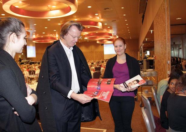 Ein Mann im Anzug zeigt einer jungen Frau ein Programmheft in einem Veranstaltungsraum.