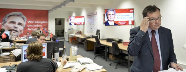Ein Mann im Anzug steht in einem Büro mit Wahlplakaten und Bildschirmen im Hintergrund.