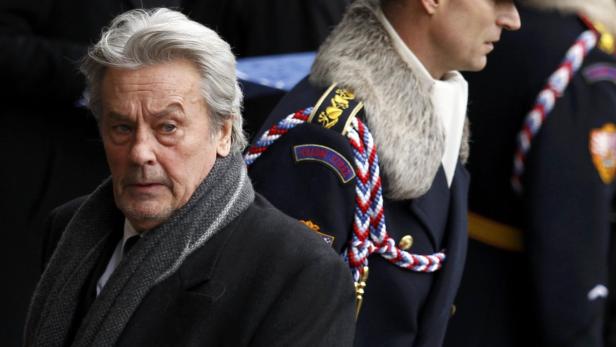 Alain Delon mit grauem Schal neben einem uniformierten Mann.