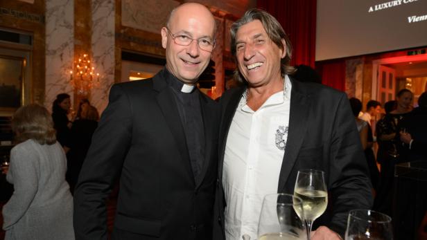 Ein Priester und ein Mann im Anzug lächeln auf einer Veranstaltung in die Kamera.
