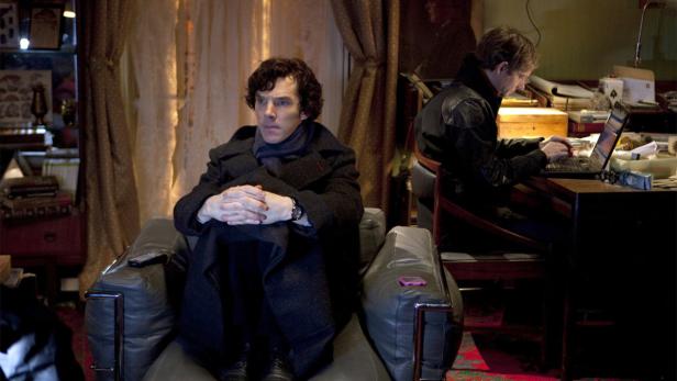 Benedict Cumberbatch als Sherlock Holmes sitzt in einem Sessel, während John Watson am Laptop arbeitet.