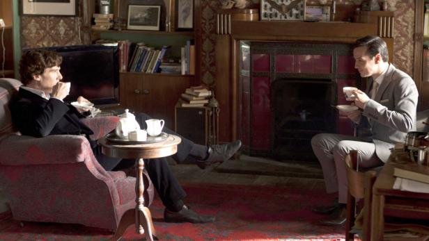 Sherlock Holmes und Moriarty trinken Tee in einem gemütlichen Wohnzimmer.