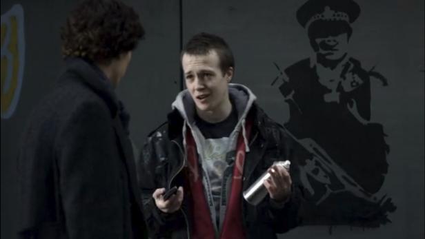 Ein junger Mann mit Spraydose steht vor einer Wand mit Graffiti, auf der ein Polizist abgebildet ist.