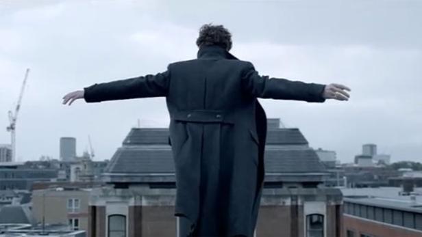 Sherlock Holmes steht mit ausgebreiteten Armen auf einem Dach in London.