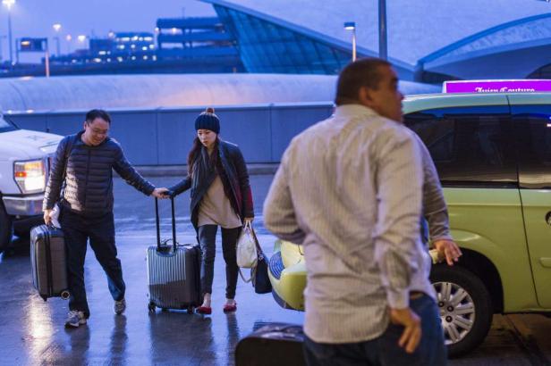 Zwei Reisende mit Gepäck verlassen ein Flughafengebäude, während ein Taxifahrer wartet.