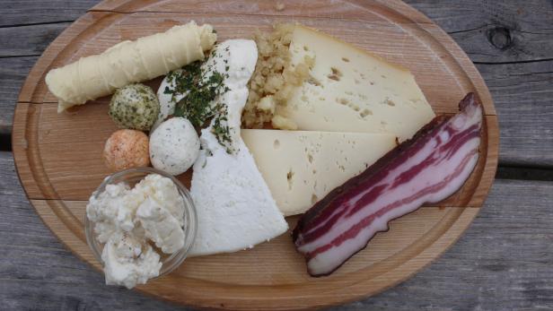 Eine Auswahl an Käse und Speck auf einem Holzbrett angerichtet.