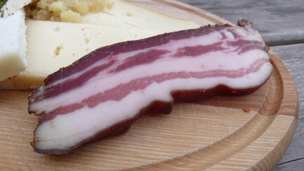 Eine Scheibe Speck liegt neben Käse auf einem Holzbrett.