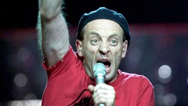 Ein Mann mit rotem T-Shirt und schwarzer Mütze singt in ein Mikrofon.