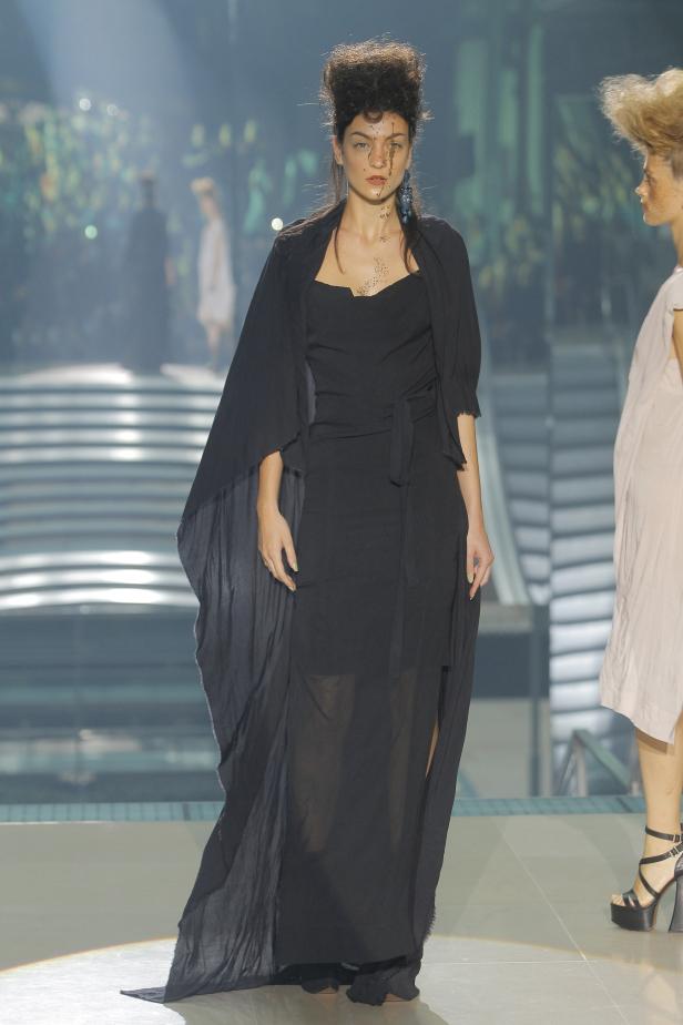 Eine Frau mit Gesichtsbemalung präsentiert ein schwarzes Kleid auf dem Laufsteg.