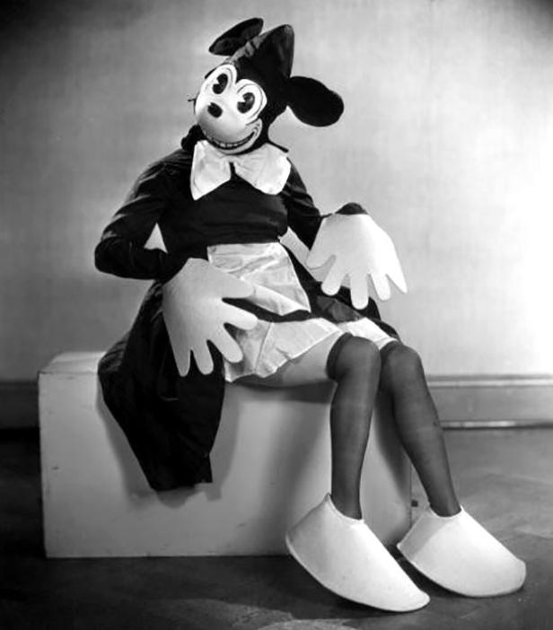 Eine Person in einem unheimlichen Mickey-Mouse-Kostüm sitzt auf einem weißen Würfel.