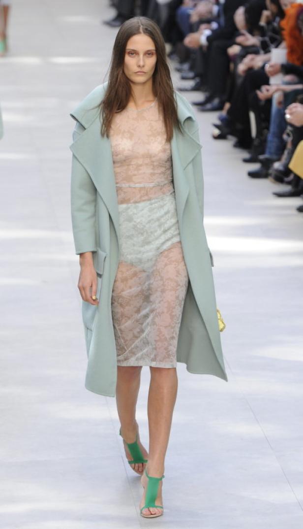 Eine Frau in einem transparenten Kleid und einem hellblauen Mantel auf einem Laufsteg.