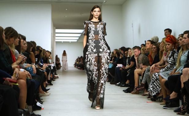 Ein Model präsentiert ein transparentes, schwarzes Kleid mit weißen Blumen auf einem Laufsteg.