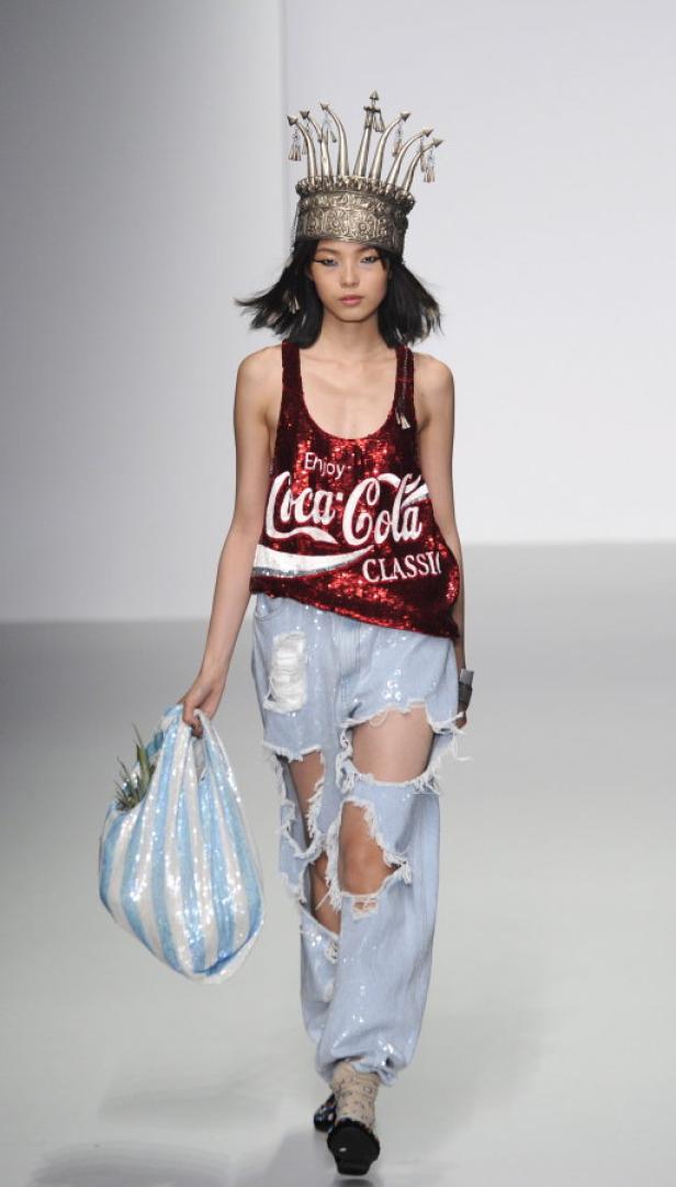 Eine Frau mit Krone und Coca-Cola-Top auf dem Laufsteg präsentiert zerrissene Jeans und eine gestreifte Tasche.