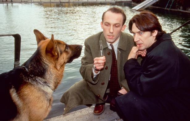 Zwei Männer und ein Schäferhund sitzen am Ufer eines Gewässers.