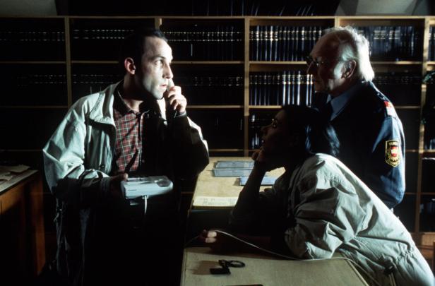 Ein Mann telefoniert, während eine Frau und ein älterer Mann mit Uniform daneben stehen.