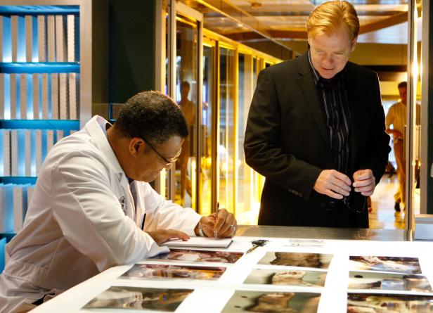 Ein Mann im Laborkittel macht Notizen, während ein anderer Mann im Anzug daneben steht.