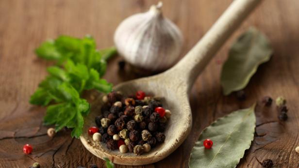 Ein Holzlöffel gefüllt mit verschiedenen Pfefferkörnern, Knoblauch, Lorbeerblatt und Petersilie auf einem Holztisch.