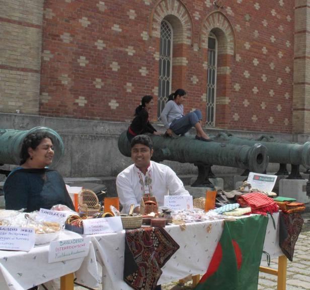 Ein Mann und eine Frau verkaufen bangladeschische Produkte an einem Stand, während zwei Frauen auf Kanonen sitzen.