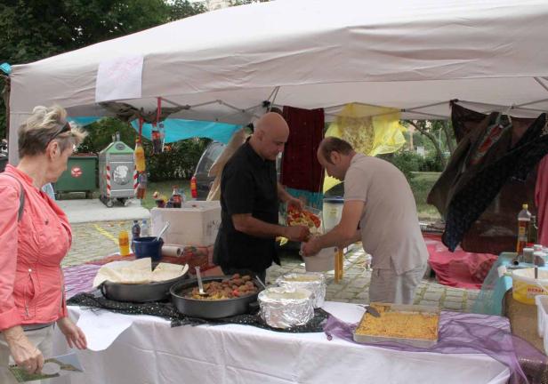 Zwei Männer bereiten Speisen an einem Stand unter einem Zelt zu, während eine Frau zusieht.