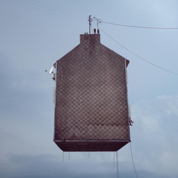 Ein Haus mit Ziegelfassade schwebt in der Luft, gehalten von dünnen Seilen.