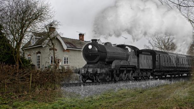 Eine alte Dampflokomotive fährt an einem Landhaus vorbei.