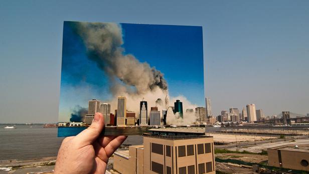 Eine Hand hält ein Foto der rauchenden Twin Towers vor der Skyline von New York.