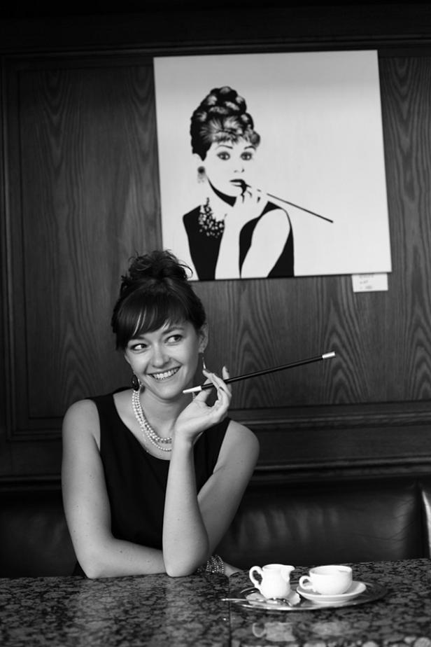 Eine Frau im Stil von Audrey Hepburn sitzt mit einer Zigarettenspitze in einem Café.