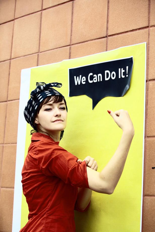 Eine Frau im Rosie-the-Riveter-Stil macht vor einem Plakat mit der Aufschrift „We Can Do It!“ eine Muskelpose.