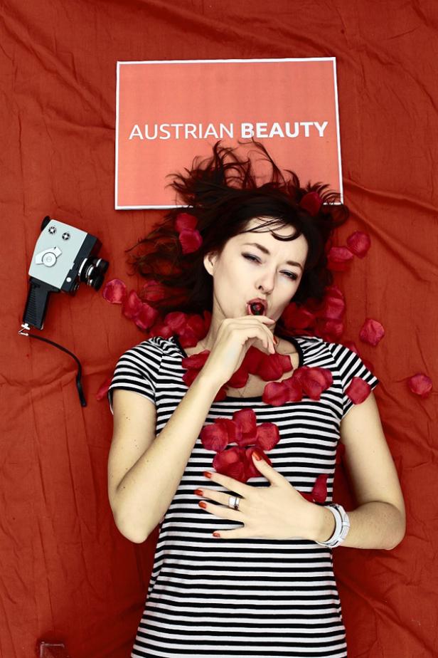 Eine Frau liegt auf einem roten Tuch mit Rosenblättern und einer Kamera neben sich, ein Schild sagt „Austrian Beauty“.