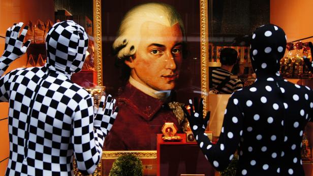 Zwei Personen in Ganzkörperanzügen betrachten ein Porträt von Mozart in einem Schaufenster.