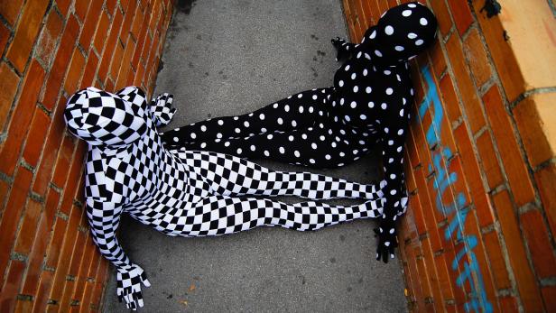 Zwei Personen in Ganzkörperanzügen mit Schachbrett- und Punktmuster liegen in einer Gasse.