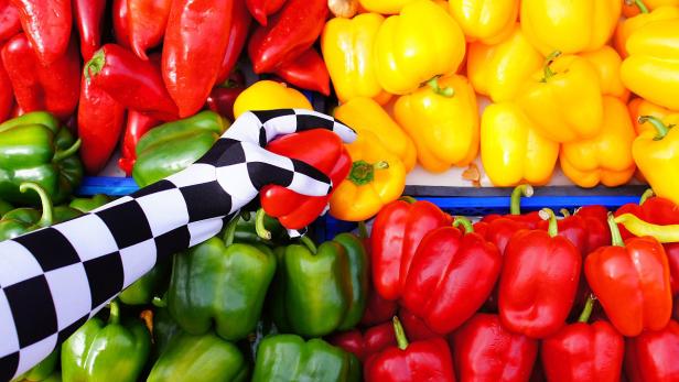 Eine Hand mit Schachbrettmuster greift nach einer roten Paprika zwischen gelben, grünen und roten Paprika.