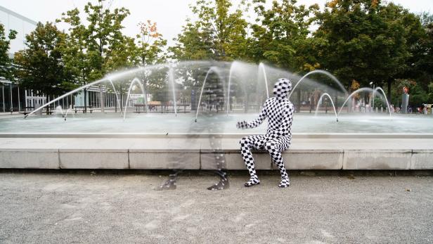 Eine Person im Schachbrettmuster-Anzug sitzt vor einem Brunnen.