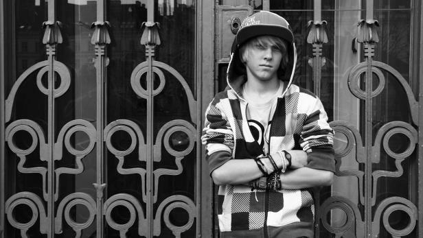 Ein junger Mann mit Kappe und karierter Jacke steht vor einem verzierten Metallgitter.