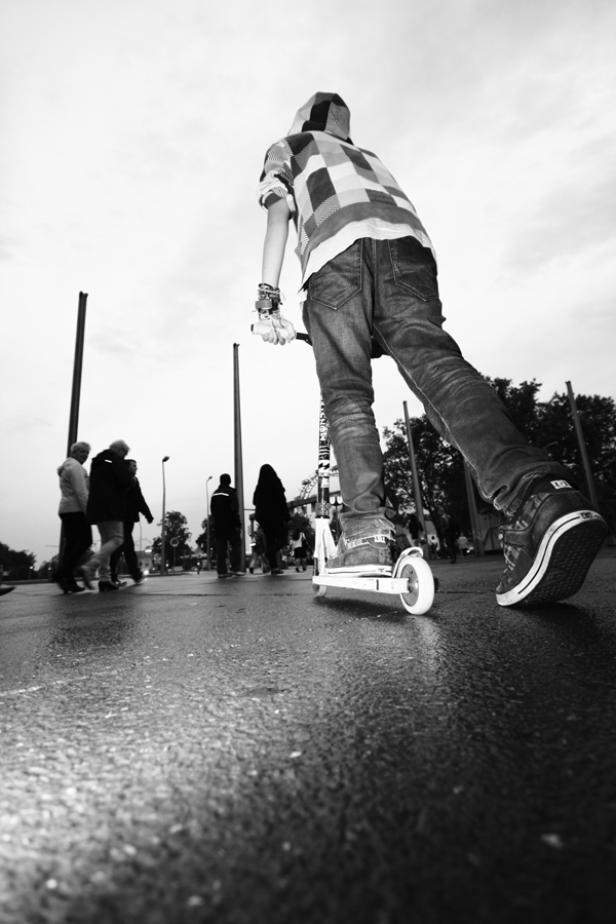 Ein Jugendlicher fährt mit einem Roller auf einer nassen Straße, im Hintergrund sind Passanten zu sehen.