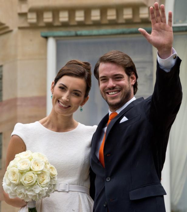 Ein strahlendes Paar, die Frau mit einem weißen Rosenstrauß, der Mann winkend, posiert vor einem Gebäude.