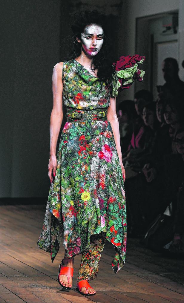 Ein Model mit weißer Gesichtsbemalung trägt ein geblümtes Kleid mit passenden Schuhen auf dem Laufsteg.