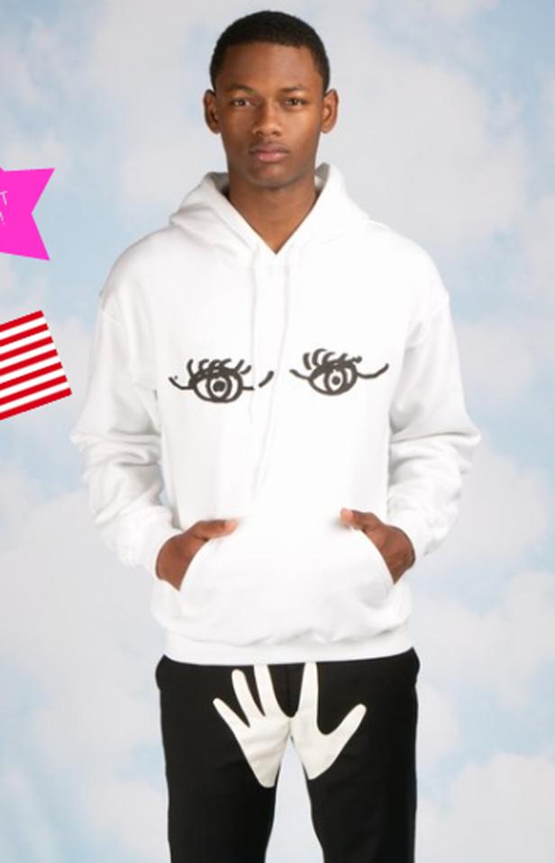 Ein Mann trägt einen weißen Hoodie mit aufgedruckten Augen und eine schwarze Hose mit einer weißen Hand darauf.